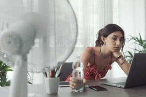 Photo of a woman cooling herself with a fan while working in an office in need of an AC duct cleaning.