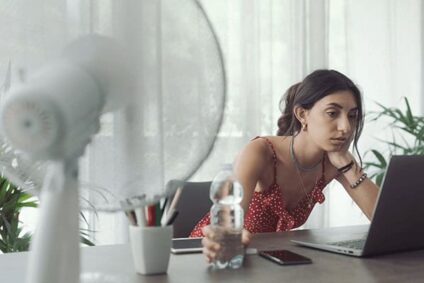 Photo of a woman cooling herself with a fan while working in an office in need of an AC duct cleaning.